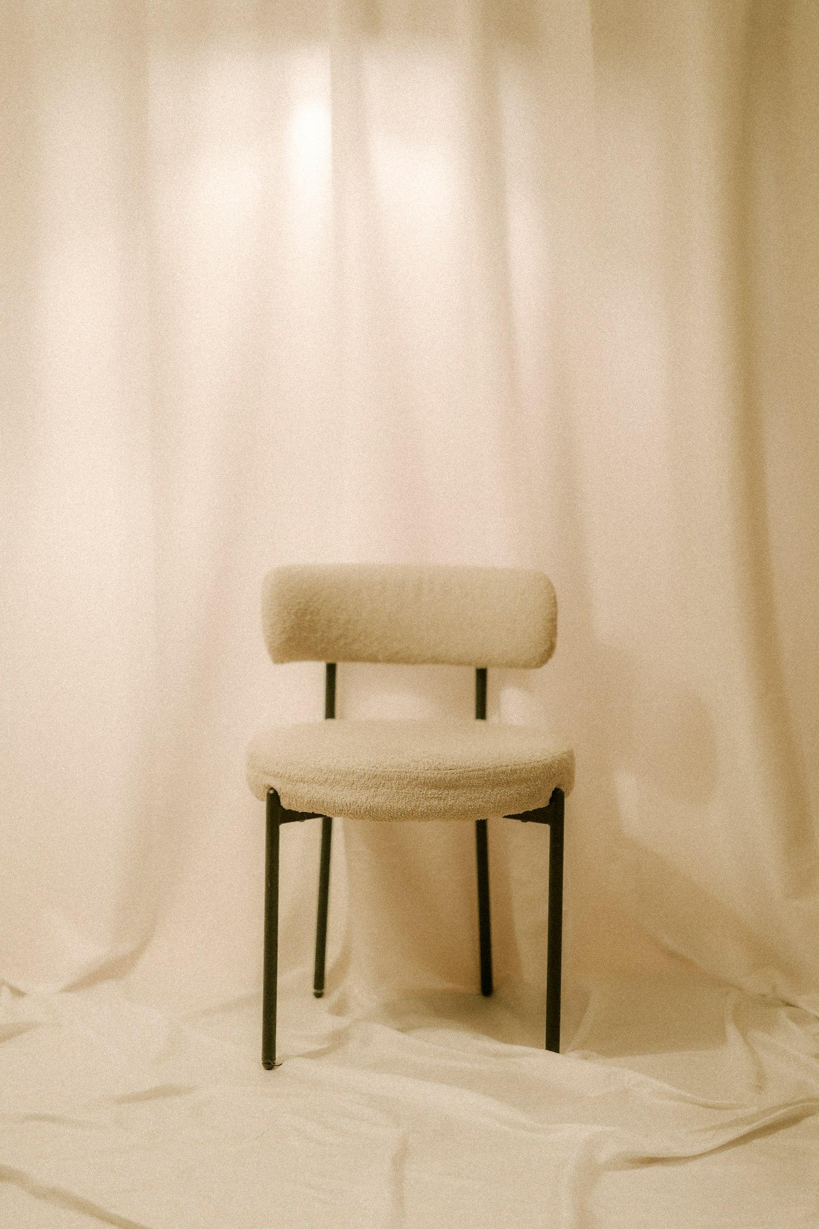 A single pale chair standing before tall white curtains.