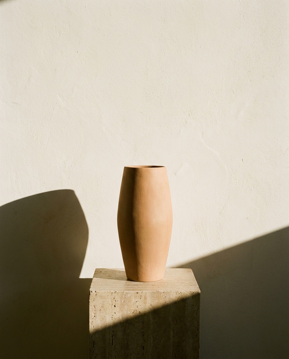 A tall unglazed terracotta vessel on a travertine plinth, cut by a hard diagonal of sun.